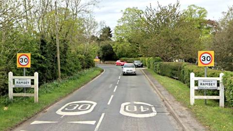 Signs on both sides of the road marking the gateway to Ramsey, accompanied by 30mph warnings. There are trees either side of the road, which bends to the left in the distance. Coming in the opposite direction is a silver car. Two other cars are parked up on the verge in the distance.
