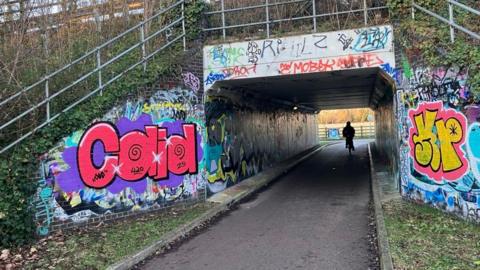 A cycle path is going under a bridge which has a road running across it. The tunnel has graffiti on it and a cyclist is coming towards the camera.