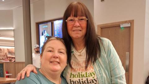 Rebecca is sat on the left of the photo and her mum Delia is stood to her right with her arm around her. Rebecca is wearing a blue t-shirt. Delia is wearing glasses and a stripy shirt over a white graphic t-shirt which says 'holiday' four times.