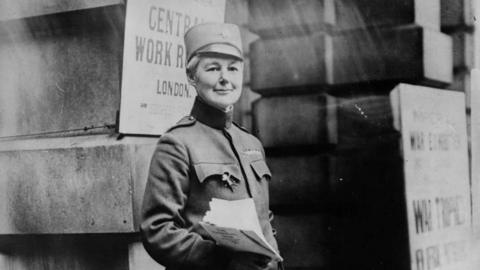 A black and white image of Flora Sandes wearing her army uniform. She is looking at the camera and smiling while she holds several documents in her hand. She is standing outside a building which has several white boards placed on it.