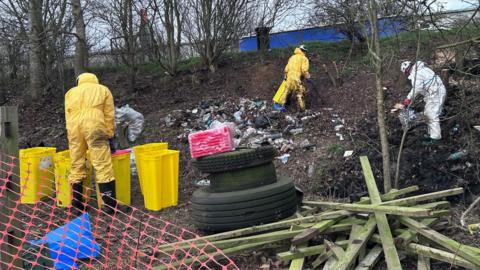 Workers wearing either white or yellow jumpsuits work to clear mess dumped on a muddy verge, behind which is the A14. The mess includes wooden beams, general litter and tyres. A series of yellow bins are being used to store some of the rubbish.