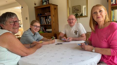 Four people are playing cards at a round table with a tablecloth. Left to right, an older woman in a light green top is next to a teenage girl wearing a stripy short-sleeved shirt, next to her is a elderly bald man in a grey T-shirt and then there is a middle-aged woman with fair hair wearing a pink top.