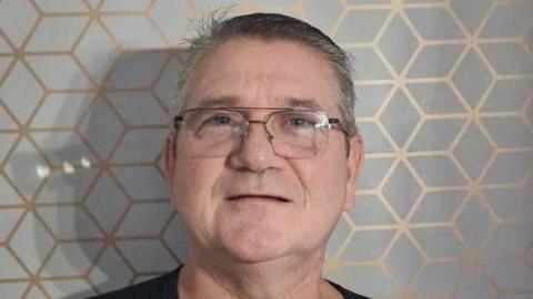 A man with short grey hair stands in front of a grey and gold patterned wallpaper. He has large glasses and is clean shaven. 