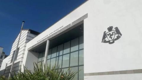 The outside of the courts of justice, which is a large, white, glass-fronted building with the government crest on the right.