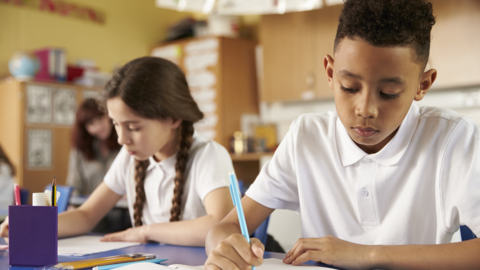 Two children sit at their school desks and write on paper. 