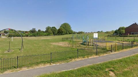 A public park on a sunny day, with outdoor play equipment including swings and slides in shot. A narrow footpath runs alongside the playground, with an iron fence following its route. Houses and trees can be seen in the background. 