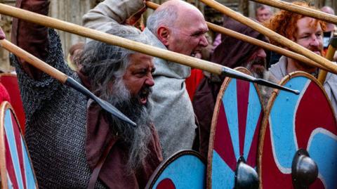 A group of men dressed as Vikings with angry expressions on their faces hold spears and shields as they look to charge at the enemy out of shot. 