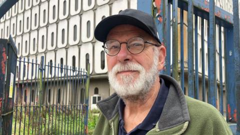John stands smiling outside Marlborough House, he is wearing a black cap and has a white beard. He is also wearing black framed glasses and a green fleece coat. "