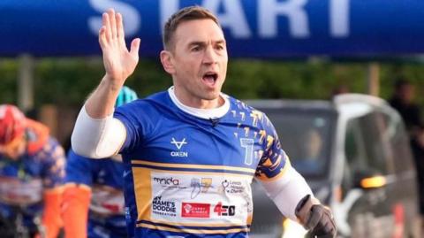 Kevin Sinfield wearing a blue and white race jersey with sponsor logos raises one hand near the starting line. The background features a large blue inflatable arch with the word ‘START’ in bold white letters.
