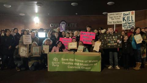 Crowds of protestors stand outside a red-brick building in the dark holding up various placards. A large green banner in the middle from the Green Party says "Save the Prince of Wales Theatre and Museum of Cannock Chase."