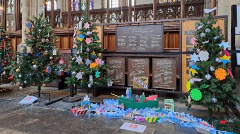 A church area with three Christmas trees that are decorated with paper decorations. There are many colourful Christmas crafts on the floor between the Christmas trees. 