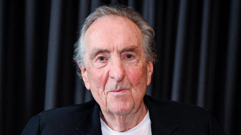 Eric Idle smiles for a photo in front of a dark curtain backdrop. He is wearing a black blazer over a white shirt.