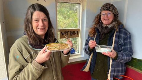 Sarah Worne in a khaki coloured jacket holds a paint dish and smiles at the camera. There is a sign behind her in a window saying wet paint. Another Helen Nicholls  woman in a blue checked jacket and leopard print hat stands to her right also holding a tub of paint.