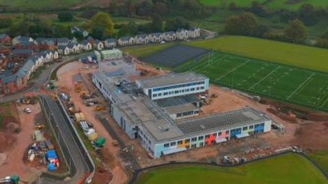 An ariel view of the Matford Brook Academy building. It is surrounded by light brown mud. It is a colourful building and has a big pitch to the right. To the right of the photo is a main road.