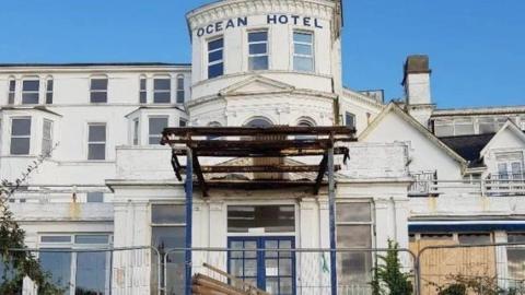 White frontage of hotel with some windows and others boarded up a metal fence stands across the front