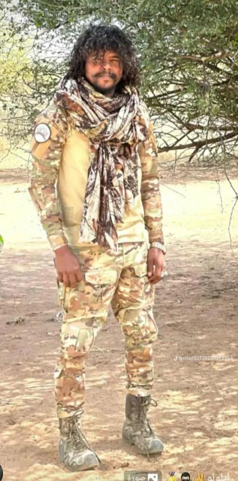 A man with dark curly hair wearing camouflage clothing standing outside next to a tree.