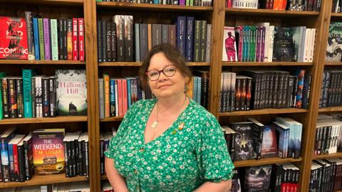 A woman with short, wavy brown hair is pictured in front of a large book case. She has brown glasses and is wearing a floral green top with buttons down the middle.