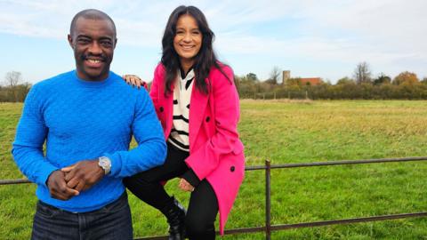 Tim Campbell stands next to BBC presenter, Sonali Shah who is sitting on a thin metal fence, there is a meadow and greenery in the background. 