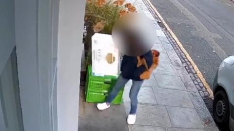 Woman in blue jeans and black coat lifts white box from behind planter and green crates. Her face is blurred. The camera shot is taken from above and shows part of the sidewalk and road.