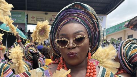 A woman in a colourful headscarf, sun glasses and a red bead necklace is holding a yellow feather fan.