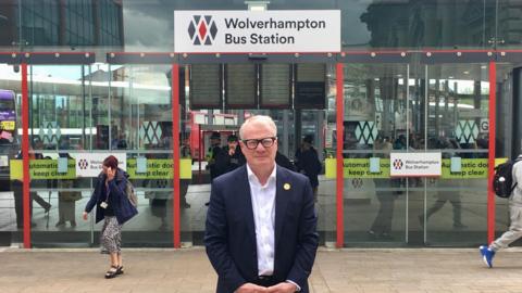 The West Midlands mayor Richard Parker, a middle-aged man with white hair and spectacles, stands in front of a set of glass doors under a sign reading "Wolverhampton Bus Station". He is wearing a blue suit jacket and a white open-necked shirt.