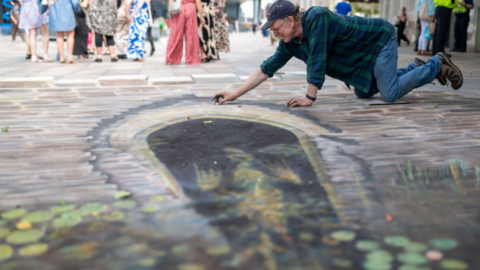 Artwork showing a water gate can be seen on the ground drawn in chalk. A man is drawing it as he kneels on the ground. He is wearing a green and blue shirt with a baseball cap on. There are people stood in groups in the background.