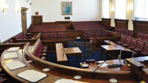 An empty Royal Court in Guernsey. It's full of red chairs and they are organised in the style of a debating chamber. The view is from the top bench. 