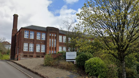 A road leading towards a red brick building with several large trees in front of it. A sign saying Oakbridge is in front of it. 