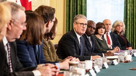 Sir Keir Starmer addresses members of the cabinet. He is wearing glasses and a dark suit and tie with a white shirt. Members of the cabinet are looking towards Sir Keir. In front of them are papers, drinks glasses and mugs.