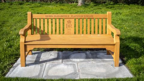 A golden bench in a park which says Queen Elizabeth II Platinum Jubilee. It also has the late Queen's emblem. 
