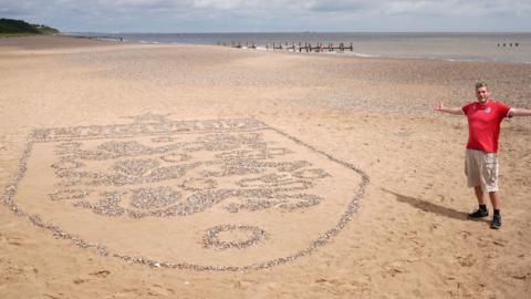 A large England crest made out of stones on a beach. Next to it is a man wearing shorts and a red England top. He has is arms out and is smiling. The sea is behind him.
