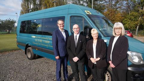 Two men and two women standing in front of a green minibus with East Hull Community Transport written on the side. The two women have black trousers, white shirts and black jackets on. One of the men has a bald head and glasses and is wearing a black suit, white shirt and grey tie. The other man has a blue suit, white open collar shirt and has short grey hair with glasses.