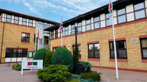 The modern building has yellow and red bricks on the ground floor and a white rendered top floor with dark grey windows. There are three flag poles on a tiled forecourt with shrubs in the middle.