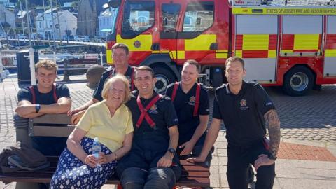 A woman posing with five firefighters on a bench, with the harbour and a fire truck in the background.