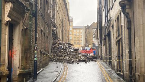 Rubble and roof timbers in a pile blocking a road with double yellow no-parking lines leading up to the scene  