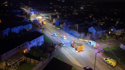A nighttime aerial view shows a residential street filled with emergency vehicles with flashing blue lights. Several police cars, ambulances, and a fire engine are positioned along the road, which is cordoned off. Houses line both sides of the street, and the wider neighbourhood is visible in the background.