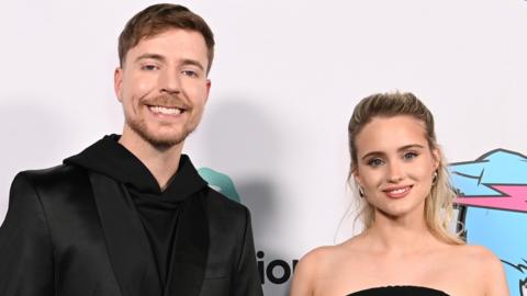 MrBeast with Thea Booysen at the premiere of Beast Games in December. MrBeast wears a black suit jacket over a black hoodie and smiles at the camera. He has a short brown beard and short brown hair. Thea wears a sleeveless black dress and has her long blonde hair styled half-up-half-down. 