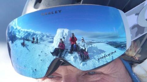 A snowy mountain landscape reflected in the mirrored ski goggles of a person wearing a helmet, showing several people sitting on the snow with wide views of peaks and sky.