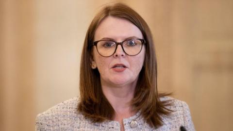 a woman with shoulder length brown hair is talking to someone off camera. She's wearing dark rimmed glasses and a light, tweed-style jacket.