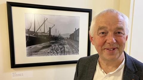 Llun o Wil Troughton yn sefyll o flaen ffotograff o'r 'Overton' - llong ager yn harbwr Aberystwyth a dynwyd ym 1926 gan Arthur Lewis.