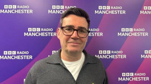 Andy Burnham, in glasses, a white T-shirt and grey jumper, stands in front of purple BBC Radio Manchester branding