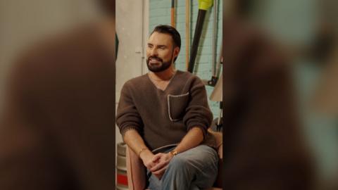 Rylan Clark has black hair and is wearing a brown jumper and grey trousers. He sits crossed-legged and smiles.