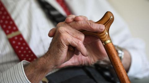 A close-up image of a man sat down, holding a walking stick - his hands are visible, as well as a brown stick, and part of his upper body. He is wearing a white shirt and red braces.