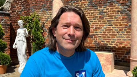 Mr Milne is smiling at the camera on a sunny day. He is sat on the flagstone stage wearing a blue top with SatT branding on (Shakespeare at the Towers). He has shoulder length brown hair. Behind him is a red bricked wall, some shrubbery and a white statue of a person holding a jug.