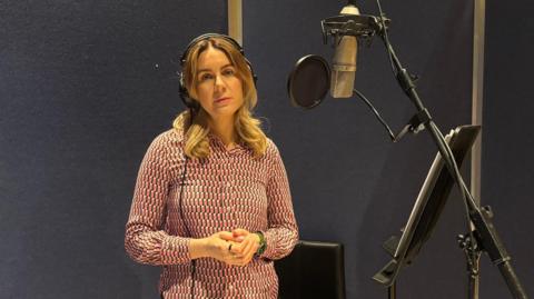 Olivia Graham stands in a recording studio in front of a mic, wearing headphones. She is wearing a geometric pattern pink and blue blouse and has her blonde/brown hair in curls around her shoulders