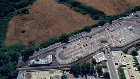 An aerial view of a road dividing an industrial campus and car park from a green area with a field and woodland.
