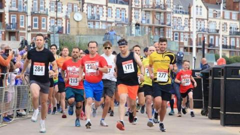 Male runners with numbers pinned to their tops head towards the photographer along a seaside promenade, with apartment buildings in the background.  Crowds stand behind barriers at the sides. One runner wears a grey Spiderman outfit.