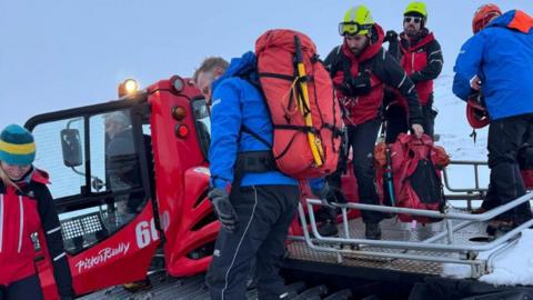 Mountain rescuers in cold weather gear and a mountain rescue vehicle in snowy conditions.