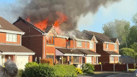 A row of semi-detached houses. A fire has broken out in one of the houses with flames and smoke coming out of the top of the roof.