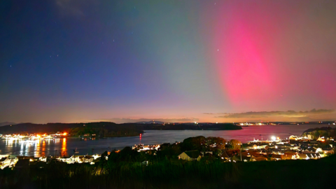 A landscape photo taken at night from a hilled area showing the Northern Lights in the sky. There are streaks of green, blue and pink lights.